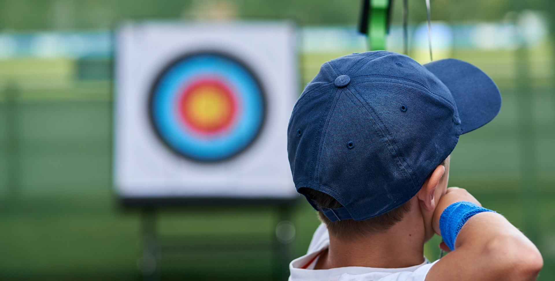 A boy in a dark-blue baseball cap, drawing a bow and aiming an arrow at a bullseye target. The image shows the boy's head and shoulders from the back. The boy is in focus and the trees and target are blurred.