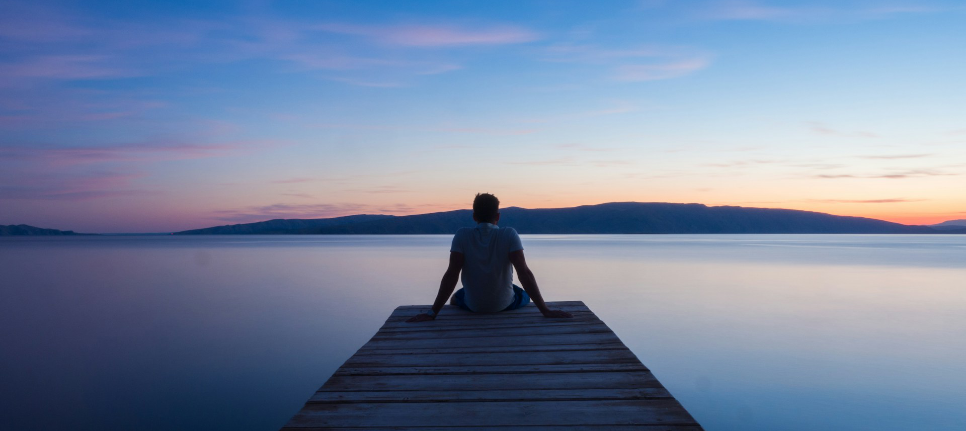 Rear solhouette of a man sitting at the end of a wooden dock. The man is leaning back on his hands and satring out at a very calm lake at sunset.