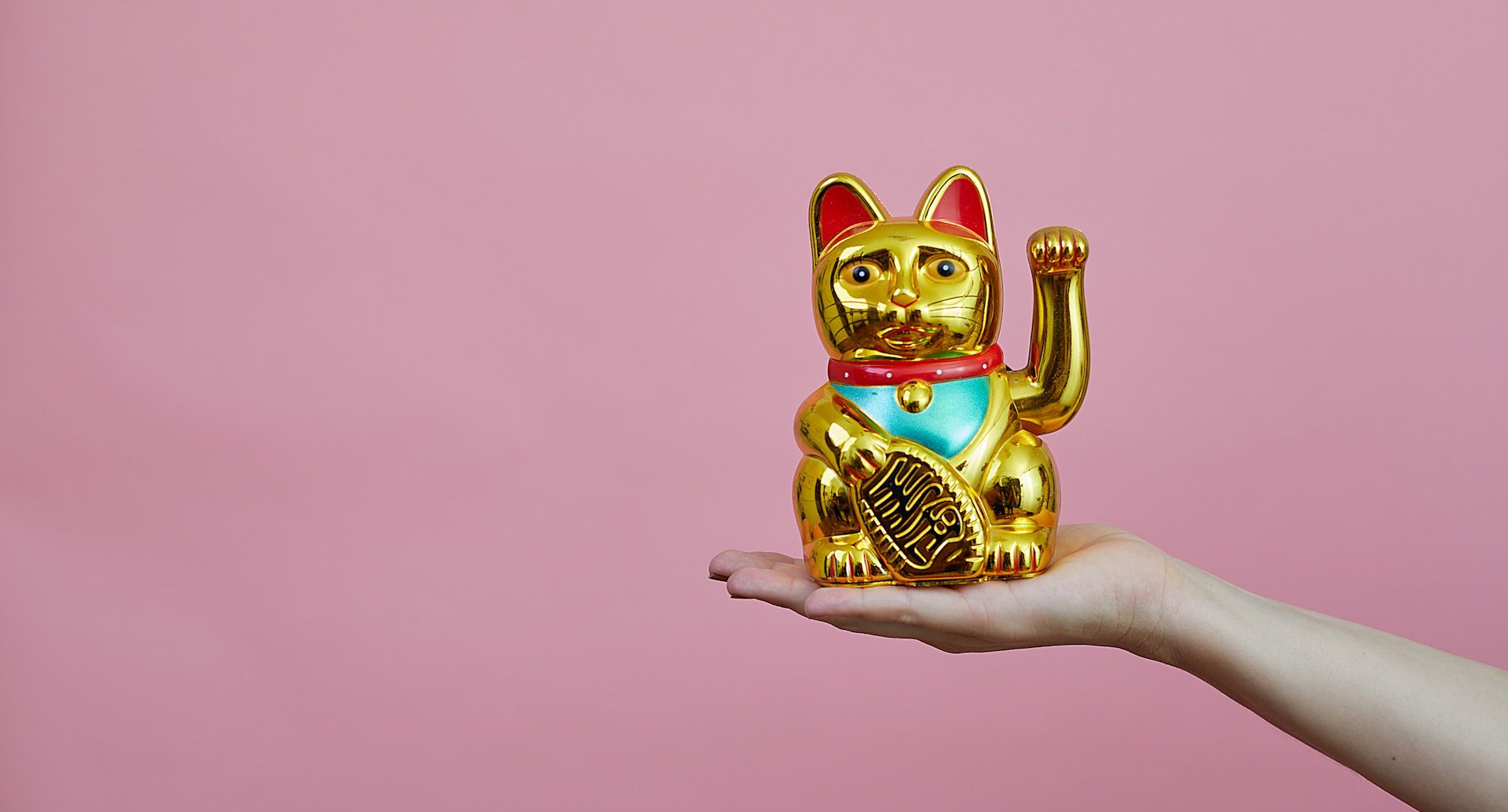 A photo of a gold maneki-neko ('beckoning cat’), a traditional Japanese figurine believed to bring its owner good luck. The maneki-neko is sitting flat on a person’s extended hand and is facing directly into the camera. The figurine's left paw is raised beside its ear. The background of the photo is a solid pink surface.